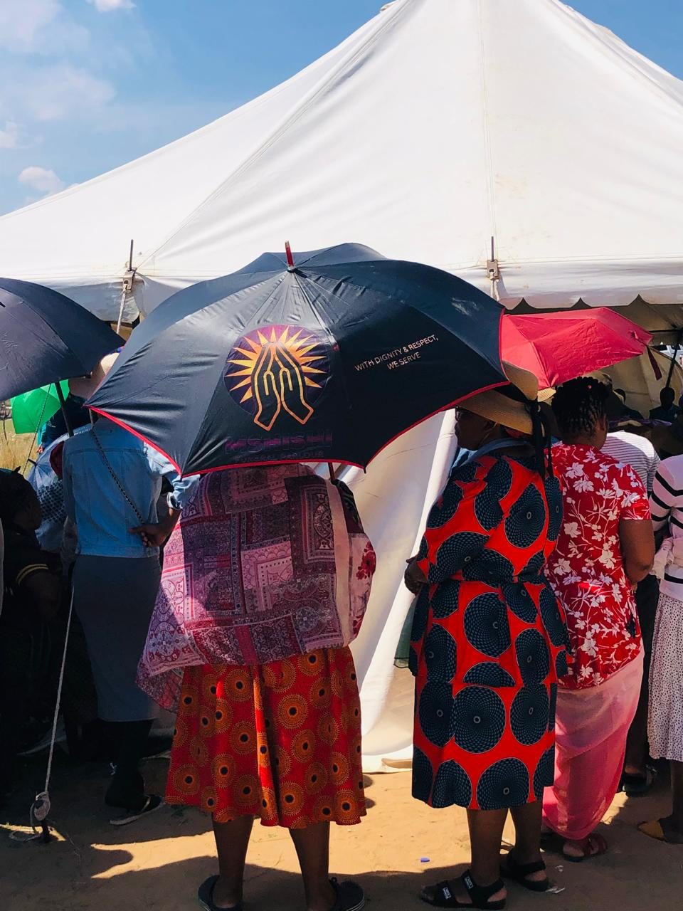 Guests gathered outside a white marquee, one holding a black branded Mshishi umbrella.