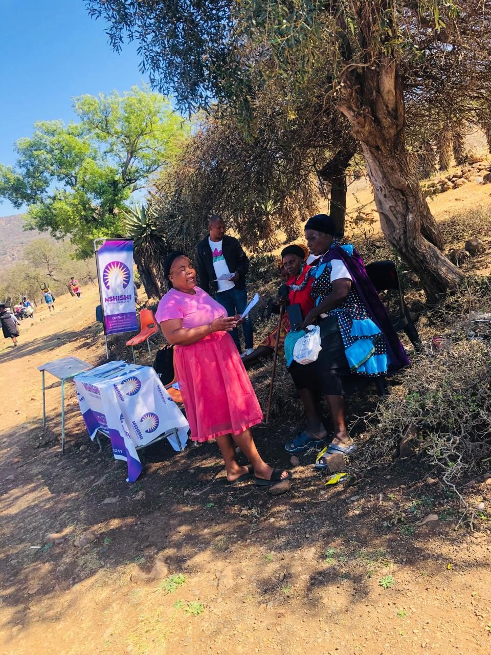 A Mshishi information table and banner stand set up under a tree while team members speak to visitors.
