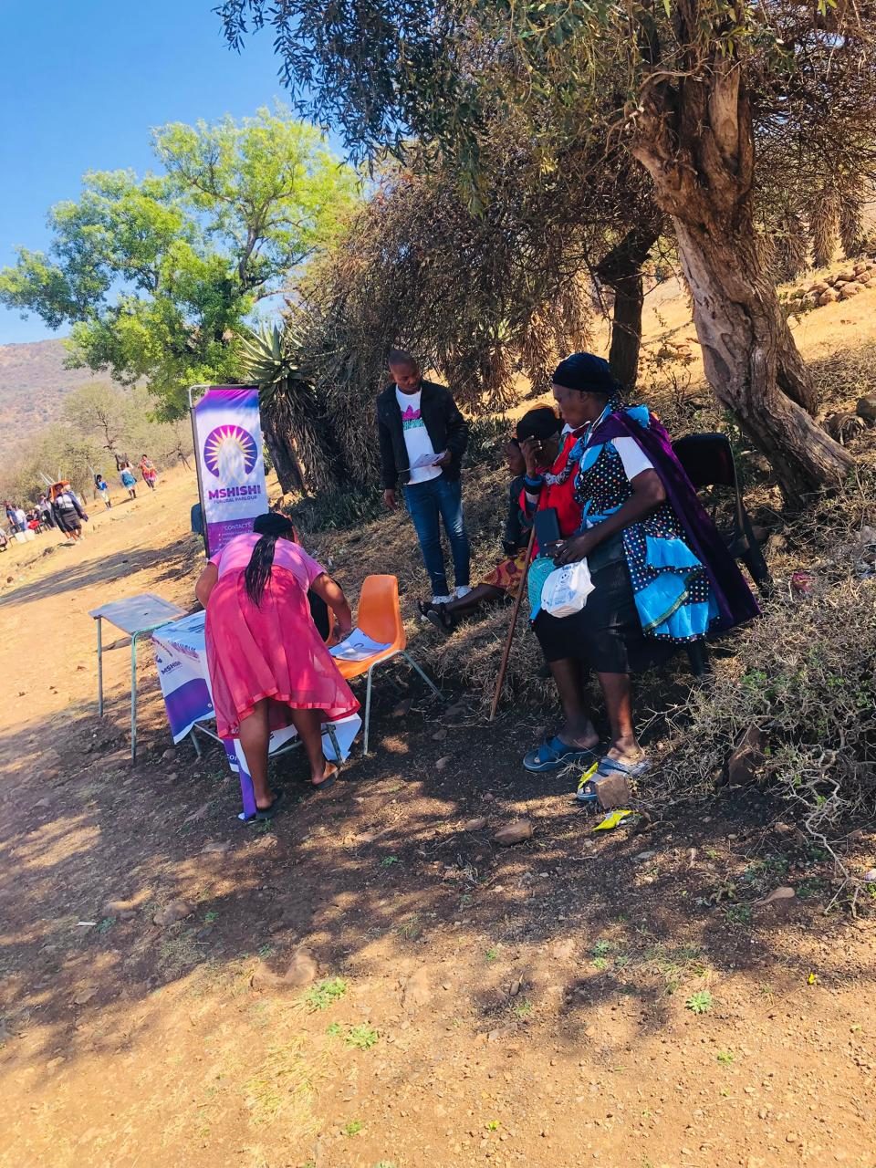 A Mshishi table and banner stand in the shade of a tree beside a dirt road, with a team member holding documents.